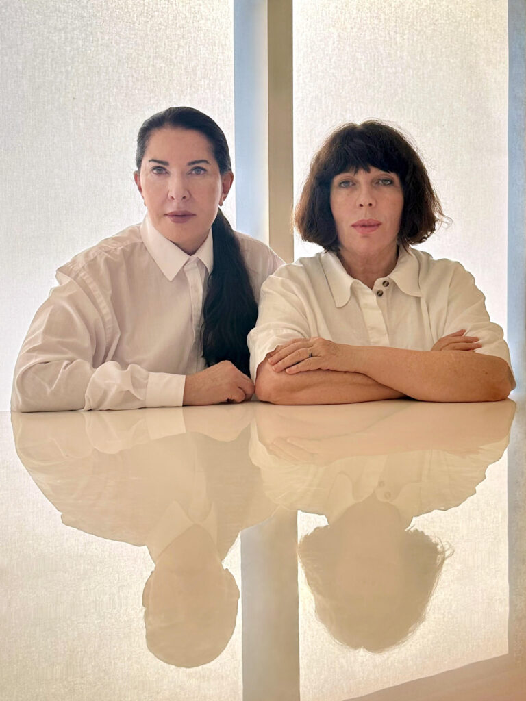 Marina Abramović and Dr. Nonna Brenner seated side by side, both in white shirts, against a softly lit backdrop, reflecting their collaborative spirit in wellness.